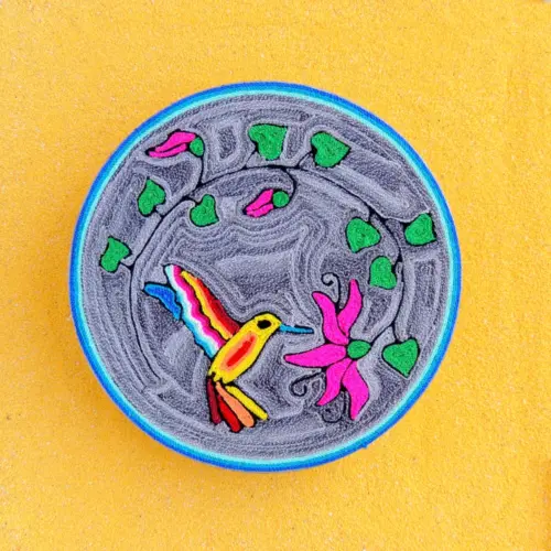 Handmade Huichol yarn painting showing a colorful hummingbird flying toward a pink flower, crafted with yarn and beeswax on a 20cm round wooden base.