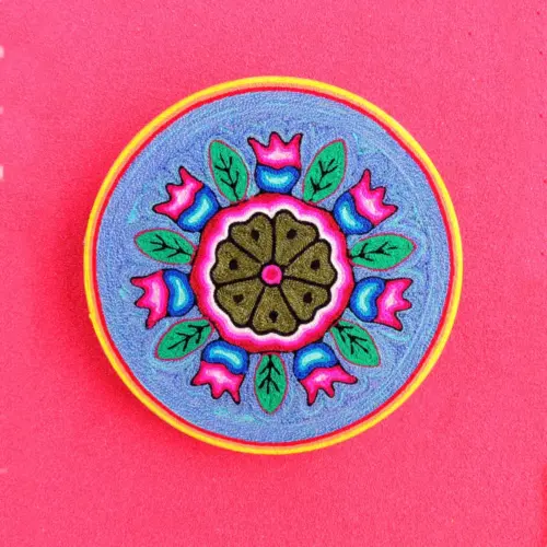 Handcrafted Huichol yarn painting of a circular peyote flower surrounded by pink petals and green leaves on a purple background.