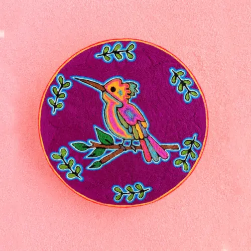 Handmade Huichol yarn painting showing a colorful hummingbird on a branch with green leaves, crafted with yarn and beeswax on a 20cm round wooden base. Front