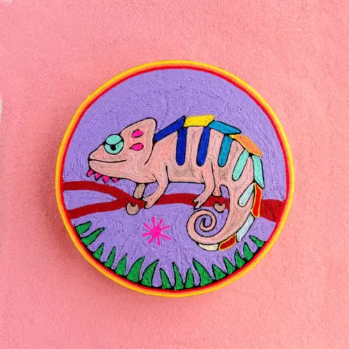 Handmade Huichol yarn painting showing a colorful chameleon resting on a branch with green grass, crafted with yarn and beeswax on a 20cm round wooden base. Front Image