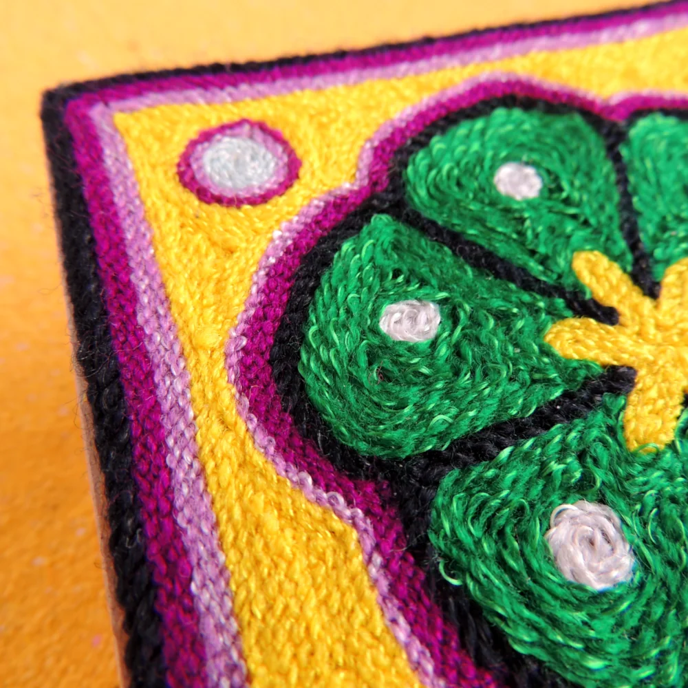 Angled corner view of a Huichol yarn painting showing yarn depth and border colors around the peyote design.