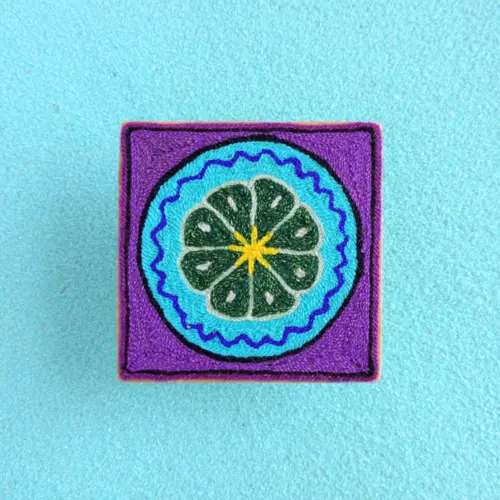 Front view of a handmade Huichol yarn painting showing a green peyote surrounded by blue and purple borders on a 10cm square wooden base.